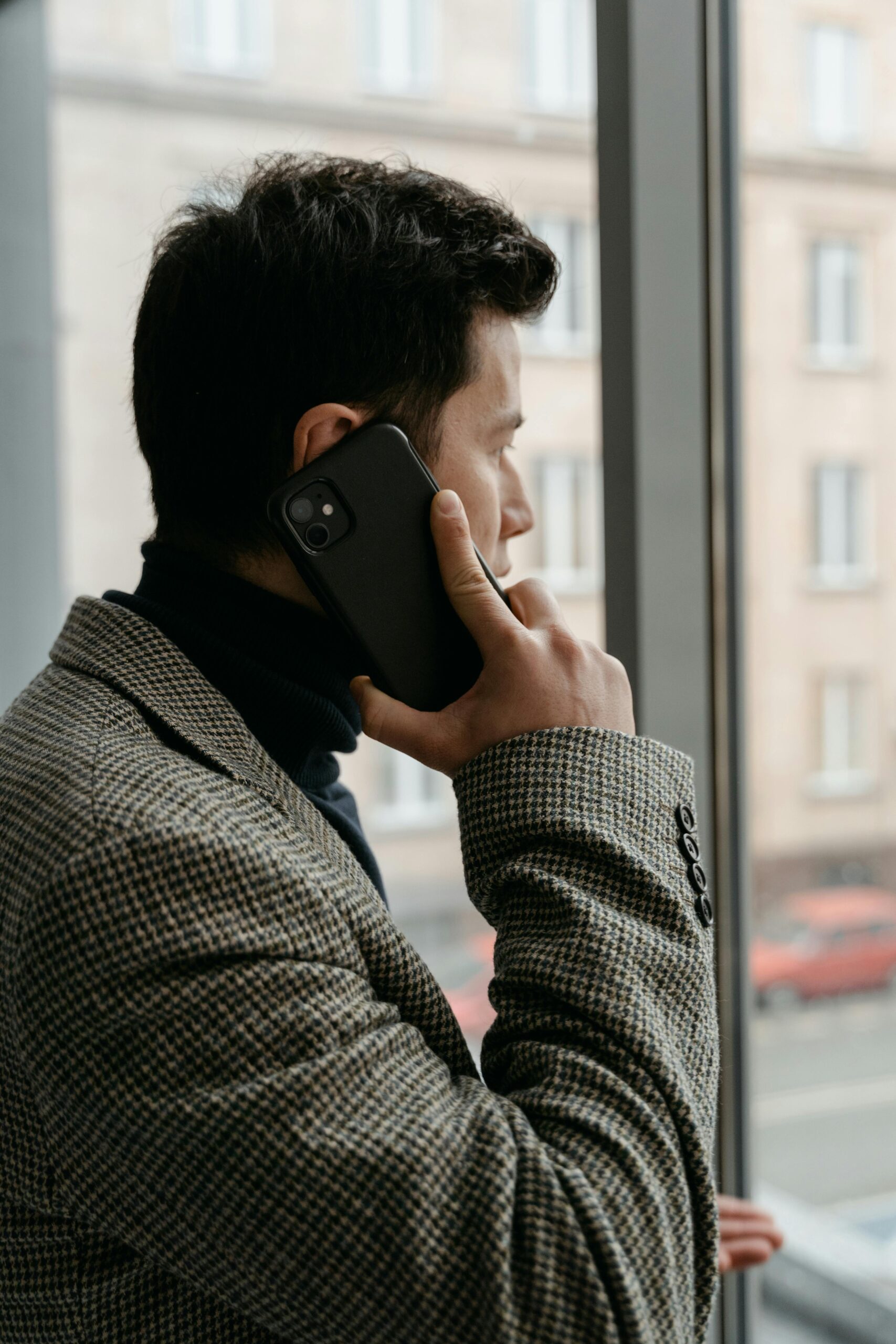 Man in a checkered blazer using a smartphone while gazing out a window.